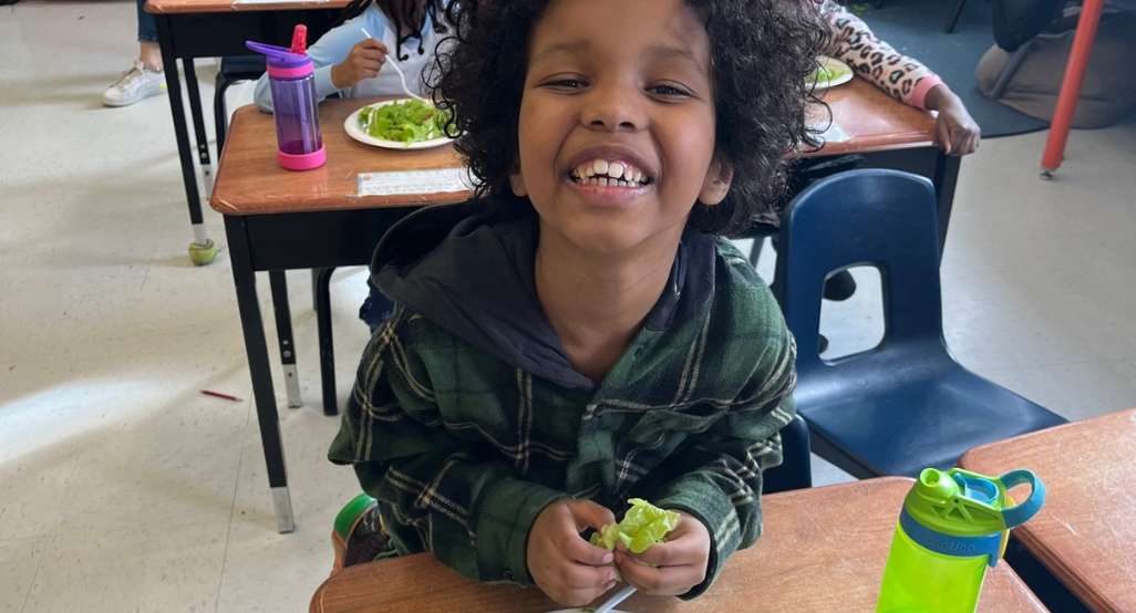 Young black child smiling into camera holding a piece of lettuce with a plate of lettuce and strawberries on the school desk in front of him
