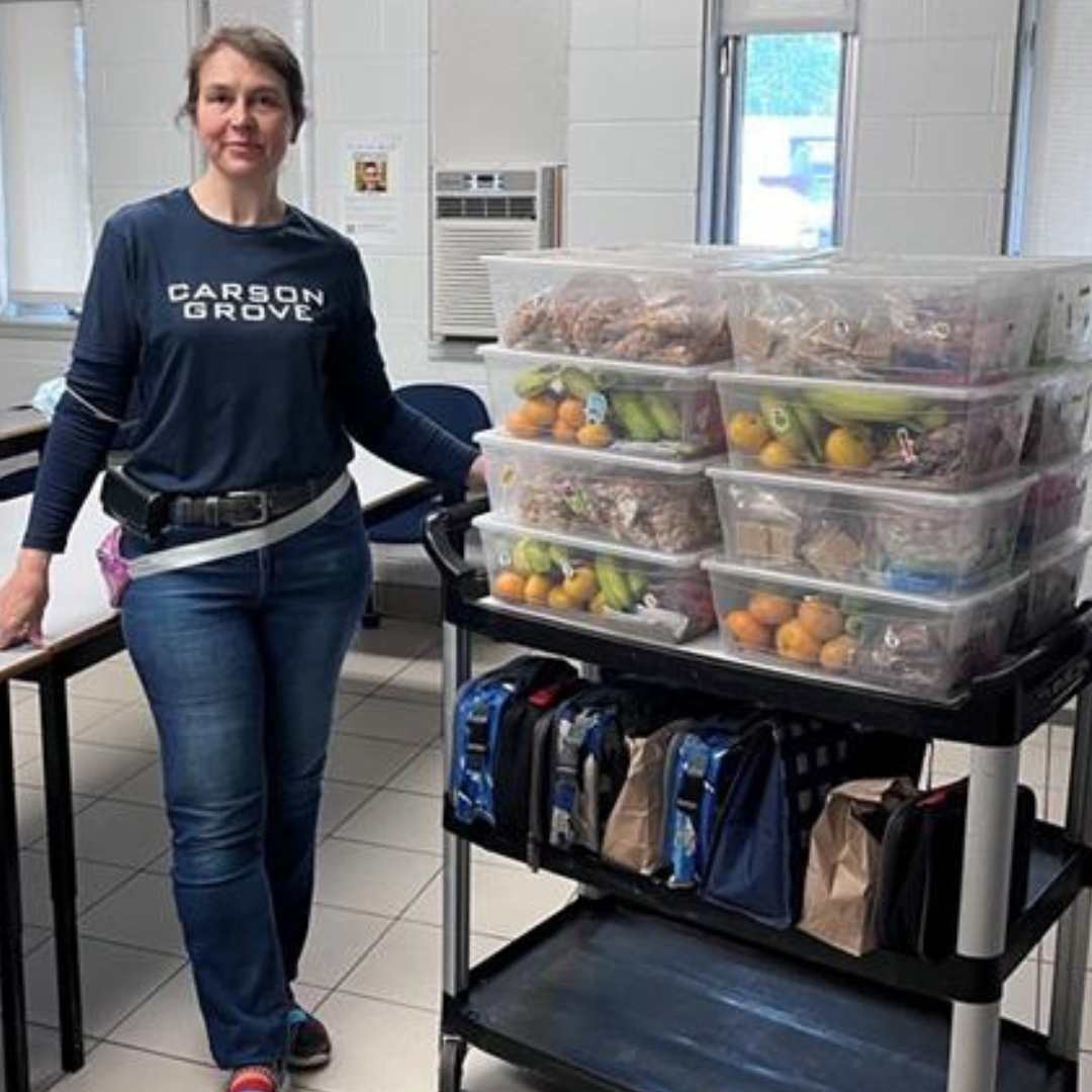 White woman wearing a blue sweatshirt and jeans looking at camera with one hand on trolley filled with bins of food and lunch bags